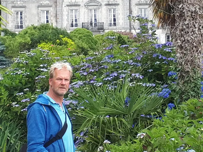 Marc Wilde in einer wundervoll angelegten grünen Gartenanlage mit Sträuchern,
 Pflanzen,
 Bäumen,
 im Hintergrund ein Herrenhaus